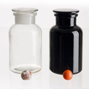 May include: Two glass bottles with stoppers, one clear and one black, on a white surface. A small orange and brown ball is in front of each bottle.