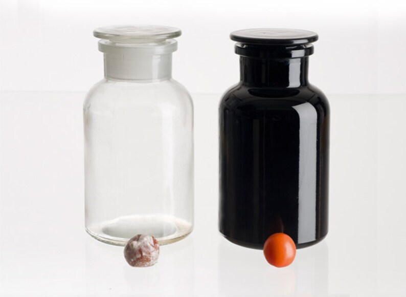 May include: Two glass bottles with stoppers, one clear and one black, on a white surface. A small orange and brown ball is in front of each bottle.