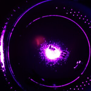 May include: A close-up of a purple and blue light source in a dark background. The light source is in the center of the image and is surrounded by a ring of smaller lights.