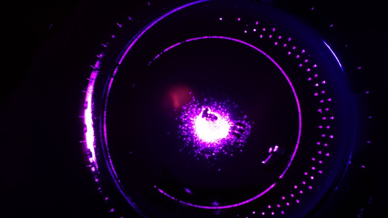 May include: A close-up of a purple and blue light source in a dark background. The light source is in the center of the image and is surrounded by a ring of smaller lights.
