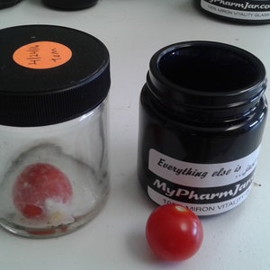 May include: Two glass jars with black lids. One jar contains a red, round object. The other jar has a black label that reads "Everything else is junk... MyPharmJar.com 100% MIRON VITALITY GLASS". A red cherry sits in front of the jars.