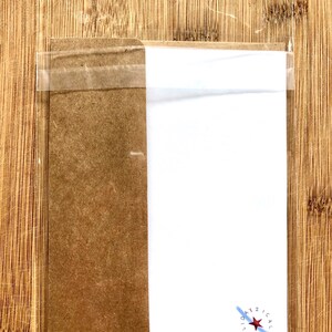 以下が含まれることがあります： 透明なプラスチック製のスリーブに入ったグリーティングカードで、茶色の段ボールの裏面にテープで留められています。カードは白で、赤い星と青色の「DAYZICAL HOLIOH」の文字が書かれています。背景は木製の表面です。