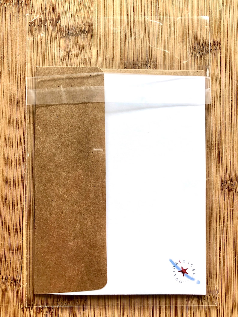 以下が含まれることがあります： 透明なプラスチック製のスリーブに入ったグリーティングカードで、茶色の段ボールの裏面にテープで留められています。カードは白で、赤い星と青色の「DAYZICAL HOLIOH」の文字が書かれています。背景は木製の表面です。