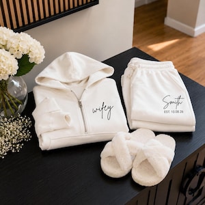 May include: A cream-colored zip-up hoodie with the word wifey printed in black script, matching sweatpants with Smith and EST. 10.08.26 printed in black, and a pair of cream-colored fuzzy crisscross slippers with open toes.