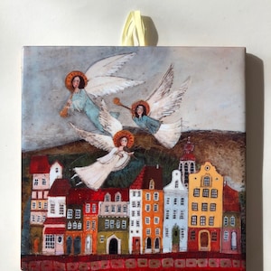 Może przedstawiać: Kwadratowy ceramiczny element artystyczny przedstawiający fantazyjną ilustrację aniołów lecących nad kolorowym miastem. Anioły, z aureolami, grają na trąbkach. Budynki pomalowane są w różnych kolorach, z czerwonymi, pomarańczowymi i żółtymi dachami.