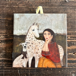 Op de afbeelding: Een schilderij van een vrouw met lang bruin haar die met een wit paard en een witte hond zit. De vrouw draagt een rode trui en een gele rok. Het paard en de hond zijn allebei gevlekt.