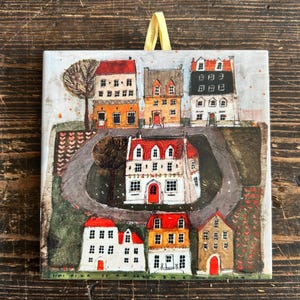 May include: Square ceramic tile with a colourful illustration of houses with red, orange, and white roofs. The houses are in a village scene with a winding road and trees. A yellow ribbon is attached for hanging.