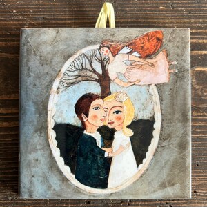 May include: A ceramic tile featuring a painted image of a couple embracing within an oval frame. Above them, an angel with red wings and a pink dress floats in the sky. The background is a watercolour wash of blue and grey.