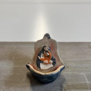 Może przedstawiać: Ceramiczna kropielnica z rzeźbionym reliefem przedstawiającym postać w ciemnoniebieskim nakryciu głowy w kropki, trzymającą mniejszą postać w pomarańczowym ubraniu. Misa w kształcie muszli ma ciemnoniebieską zewnętrzną stronę i jasne wnętrze.