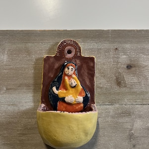 Może przedstawiać: Ceramiczna kropielnica ścienna z płaskorzeźbą postaci w ciemnoniebieskim nakryciu głowy, trzymającej dziecko w żółtym ubraniu. Tło jest brązowe z okrągłym otworem u góry, a misa ma kolor bladożółty.