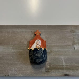Może przedstawiać: Ceramiczne kropielniczka ścienna z ciemnoniebieską, falistą miseczką i złoconym brzegiem. Tylna płyta ma czerwonobrązowy kolor i przedstawia namalowaną postać w białym welonie, zwieńczoną małym krzyżykiem na szczycie.