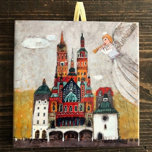 Puede incluir: Azulejo de cerámica cuadrado con una colorida ilustración de una ciudad europea con agujas y cúpulas. Un ángel con una trompeta está representado en el cielo. Una cinta amarilla está sujeta en la parte superior.
