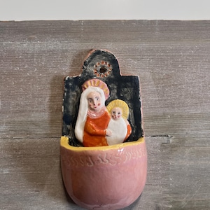 Może przedstawiać: Ceramiczna kropielnica ścienna z ręcznie malowanym reliefem przedstawiającym postać w pomarańczowej szacie trzymającą dziecko w białym otulaczu. Tło jest ciemnoniebieskie z żółtą aureolą, a misa ma delikatny różowy kolor z rzeźbionymi detalami.