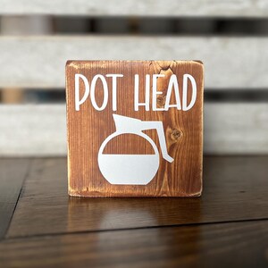 Puede incluir: Un letrero de madera con el texto "POT HEAD" en pintura blanca. Una silueta blanca de una cafetera está debajo del texto.