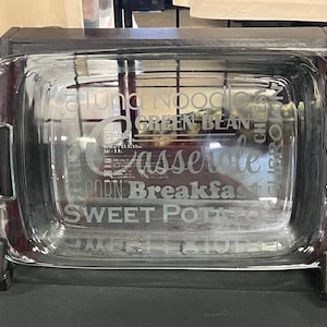 May include: A clear glass casserole dish with etched text including "Casserole", "Corn", "Breakfast", and "Sweet Potato". The rectangular dish has rounded corners and handles, and is displayed on a dark wooden stand.