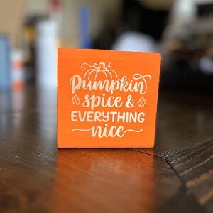 Puede incluir: Un bloque de madera naranja con las palabras "Pumpkin spice & everything nice" en escritura blanca. Un diseño de calabaza y hojas está encima de las palabras "Pumpkin spice". El bloque está sobre una superficie de madera marrón oscuro.