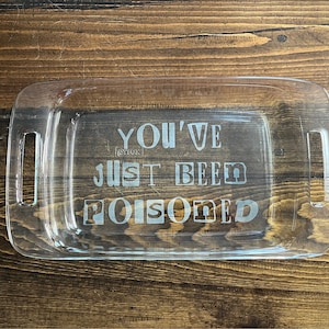Könnte beinhalten: Eine transparente Glas-Backform mit zwei Griffen. Die Form hat den Text "You've Just Been Poisoned" in den Boden geätzt.