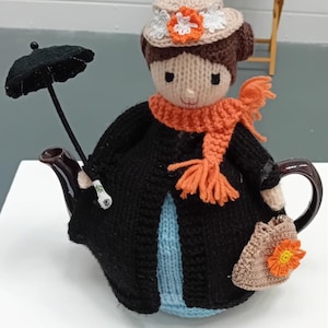 Op de afbeelding: Een gebreide theepot in de vorm van een vrouw die een zwarte jas, een oranje sjaal en een bruine hoed met een witte bloem draagt. Ze houdt een zwarte paraplu en een kleine bruine tas met een oranje bloem vast.
