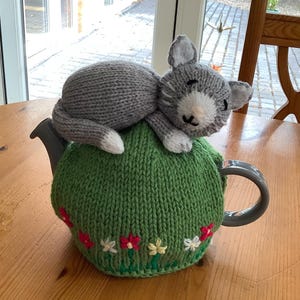 May include: A handmade, knitted tea cozy in green, adorned with embroidered flowers, topped with a gray cat. The cat has white paws and a white patch on its face. The cozy is designed to fit a teapot.