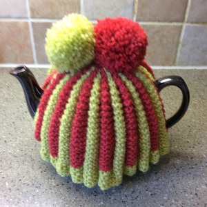 May include: A red and green striped knitted teapot cozy with two pom-poms on top. The pom-poms are green and red.