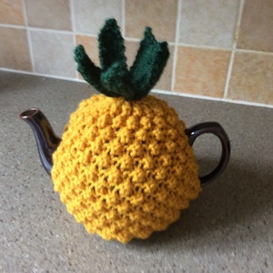 Puede incluir: Una funda de tetera de punto amarillo con forma de piña con hojas de punto verde en la parte superior.