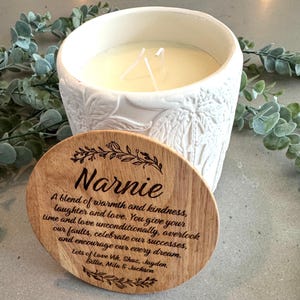 May include: A white ceramic candle holder with a textured palm leaf pattern and two wicks, accompanied by a round wooden lid engraved with the name Narnie and a heartfelt message about warmth, kindness, and love, signed by Vik, Shaz, Jayden, Billie, Mila, and Jackson.