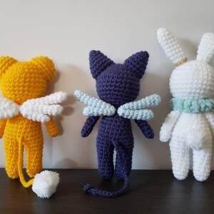 May include: Three crocheted stuffed animals: a yellow bear with white wings, a purple cat with blue wings, and a white rabbit with a light blue collar.