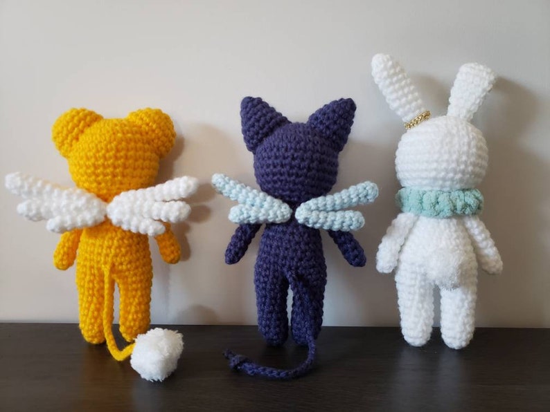 May include: Three crocheted stuffed animals: a yellow bear with white wings, a purple cat with blue wings, and a white rabbit with a light blue collar.