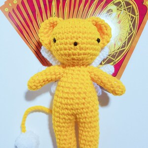 May include: A yellow crocheted plush toy with white wings and a white tail. The toy is shaped like a lion with a round head and black eyes. The toy is sitting on a white surface with a stack of red and yellow cards behind it.