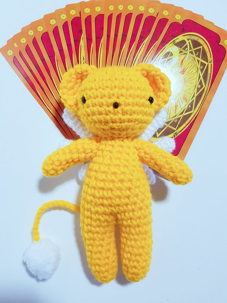 May include: A yellow crocheted plush toy with white wings and a white tail. The toy is shaped like a lion with a round head and black eyes. The toy is sitting on a white surface with a stack of red and yellow cards behind it.