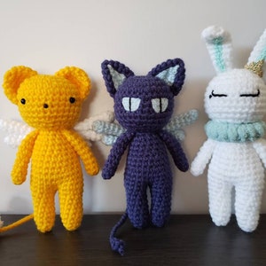 May include: Three crocheted stuffed animals: a yellow angel with white wings, a purple cat with white wings, and a white bunny with light blue ears and a light blue collar.