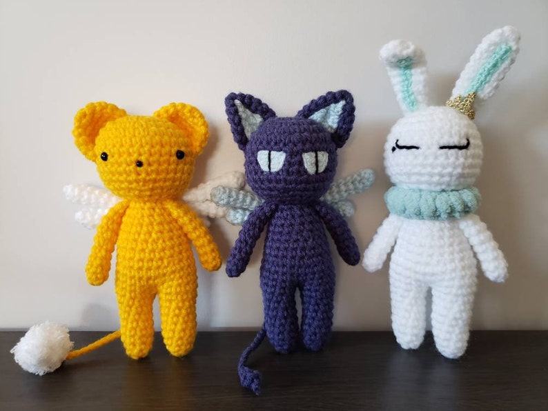 May include: Three crocheted stuffed animals: a yellow angel with white wings, a purple cat with white wings, and a white bunny with light blue ears and a light blue collar.