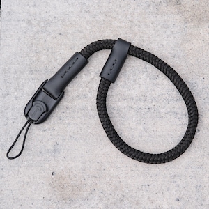 May include: Black camera wrist strap with a braided rope design. The strap features black leather accents and a quick-release mechanism. The strap is designed to provide a secure and comfortable grip for cameras.