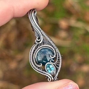 May include: A handcrafted silver pendant with a wire-wrapped design. The pendant features two blue gemstones, one larger and one smaller, set within the intricate metalwork. The pendant is held between fingers against a blurred green background.