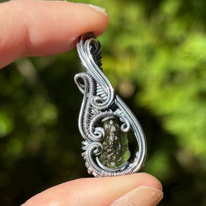 May include: A wire-wrapped pendant featuring a textured, olive-green moldavite stone encased in intricate, swirling silver-colored metal wire. The design includes a prominent bail at the top for suspension, with the metal wire forming decorative loops.