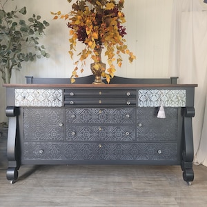 May include: A dark charcoal sideboard featuring a natural wood top, two upper drawers with a silver damask pattern, and three lower drawers with a raised embossed floral texture. The piece includes clear glass knobs and a small white tassel.