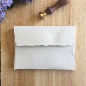 May include: A white envelope with a textured surface, lying on a wooden surface. A wooden and gold wax seal is also on the surface.