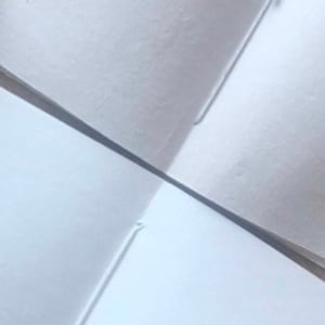 May include: Two white blank pages of a notebook, folded in half, showing the spine and the stitching along the edge.