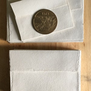 May include: A stack of white paper envelopes with a gold coin on top. The coin has a design of a bird in flight.