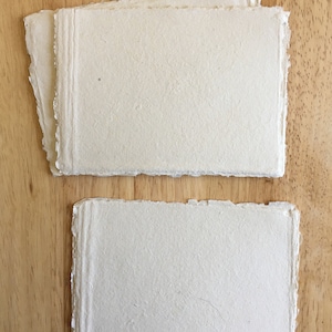 May include: Two sheets of handmade paper with a rough, textured surface. The paper is off-white in color and has a slightly irregular shape.