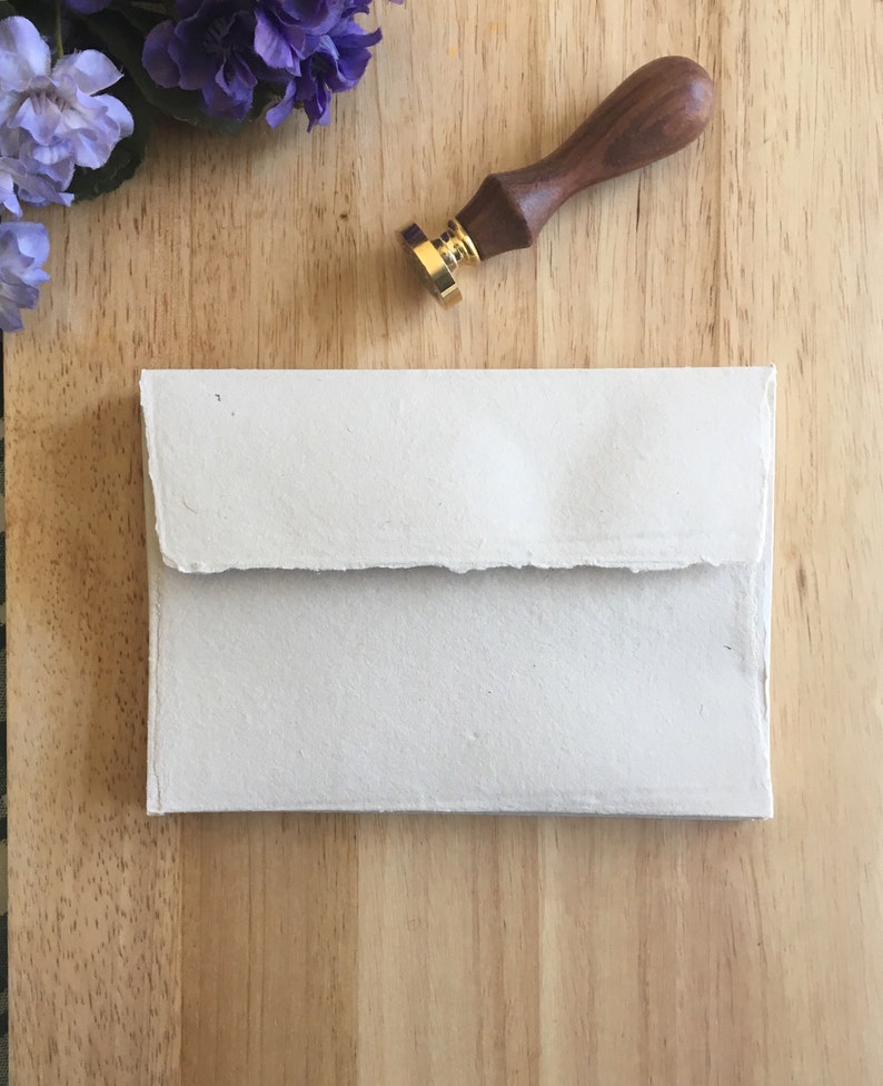 May include: A white envelope with a textured surface, lying on a wooden surface. A wooden and gold wax seal is also on the surface.