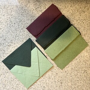 May include: Four handmade paper envelopes in different colors: burgundy, dark green, light green, and a lighter green with a speckled texture. The envelopes are arranged in a stack, with the burgundy envelope on top.