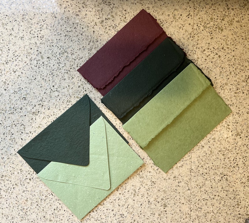 May include: Four handmade paper envelopes in different colors: burgundy, dark green, light green, and a lighter green with a speckled texture. The envelopes are arranged in a stack, with the burgundy envelope on top.