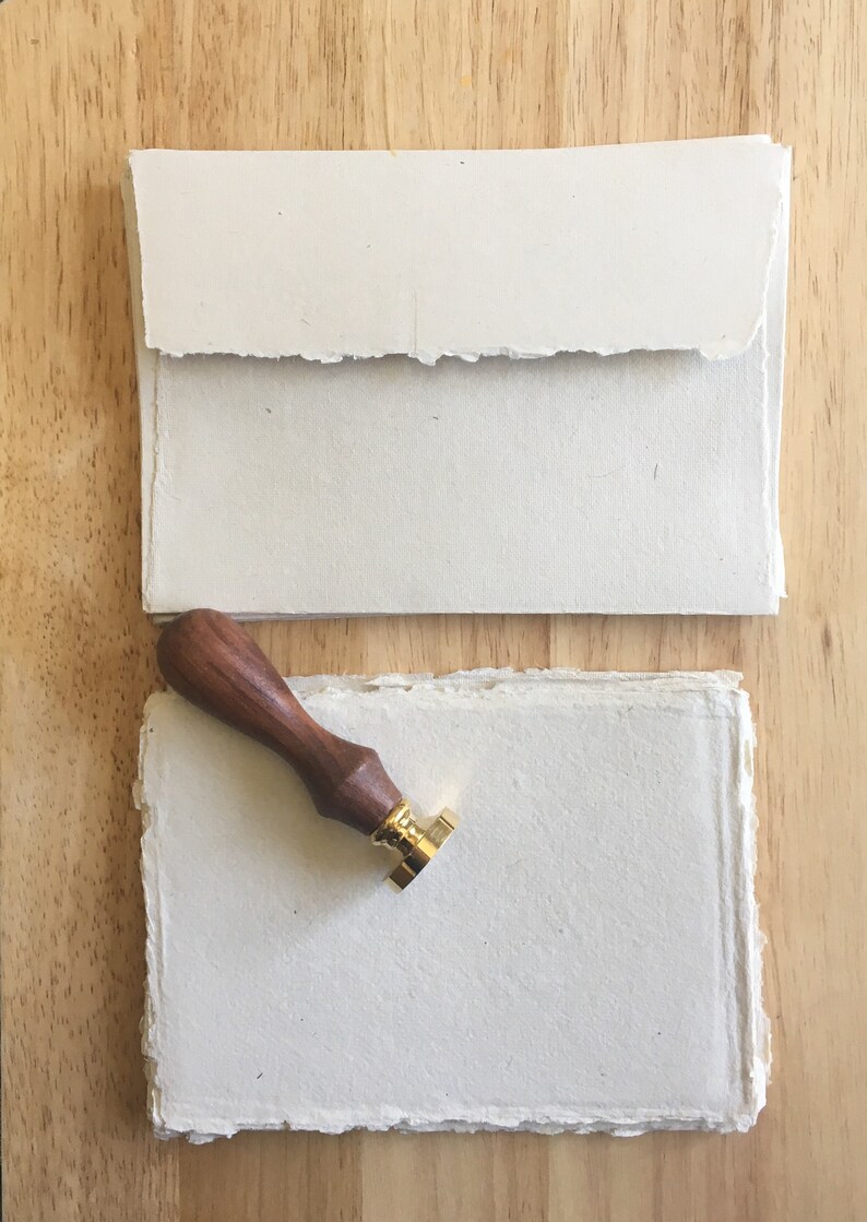 May include: A set of two blank, white, handmade paper sheets with deckled edges. A wooden stamp handle with a gold metal stamp head is on top of the sheets.