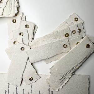 May include: A collection of rectangular off-white paper tags with deckled edges and gold-toned metal eyelets. Some tags are blank, while others feature printed black text listing menu items such as spanakopita, keftedes, and horiatiki.