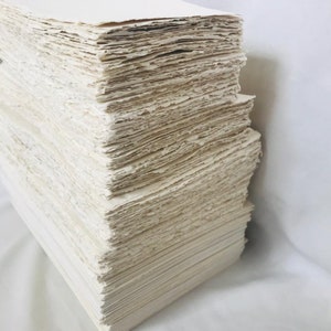 May include: A stack of handmade paper sheets, with a rough, textured surface. The paper is a light cream color and is stacked neatly.
