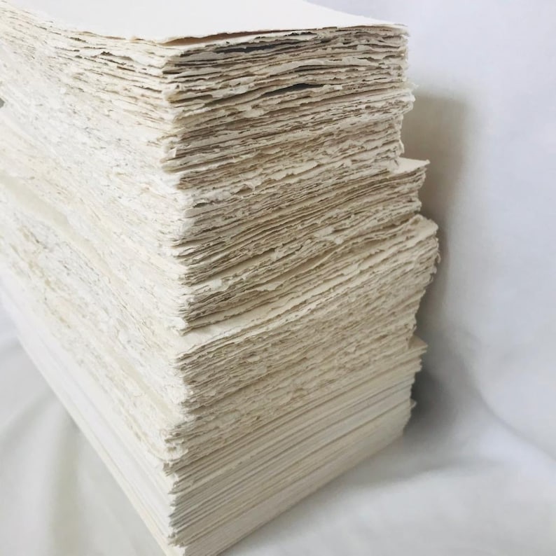 May include: A stack of handmade paper sheets, with a rough, textured surface. The paper is a light cream color and is stacked neatly.