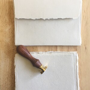 May include: A set of two blank, white, handmade paper sheets with deckled edges. A wooden stamp handle with a gold metal stamp head is on top of the sheets.