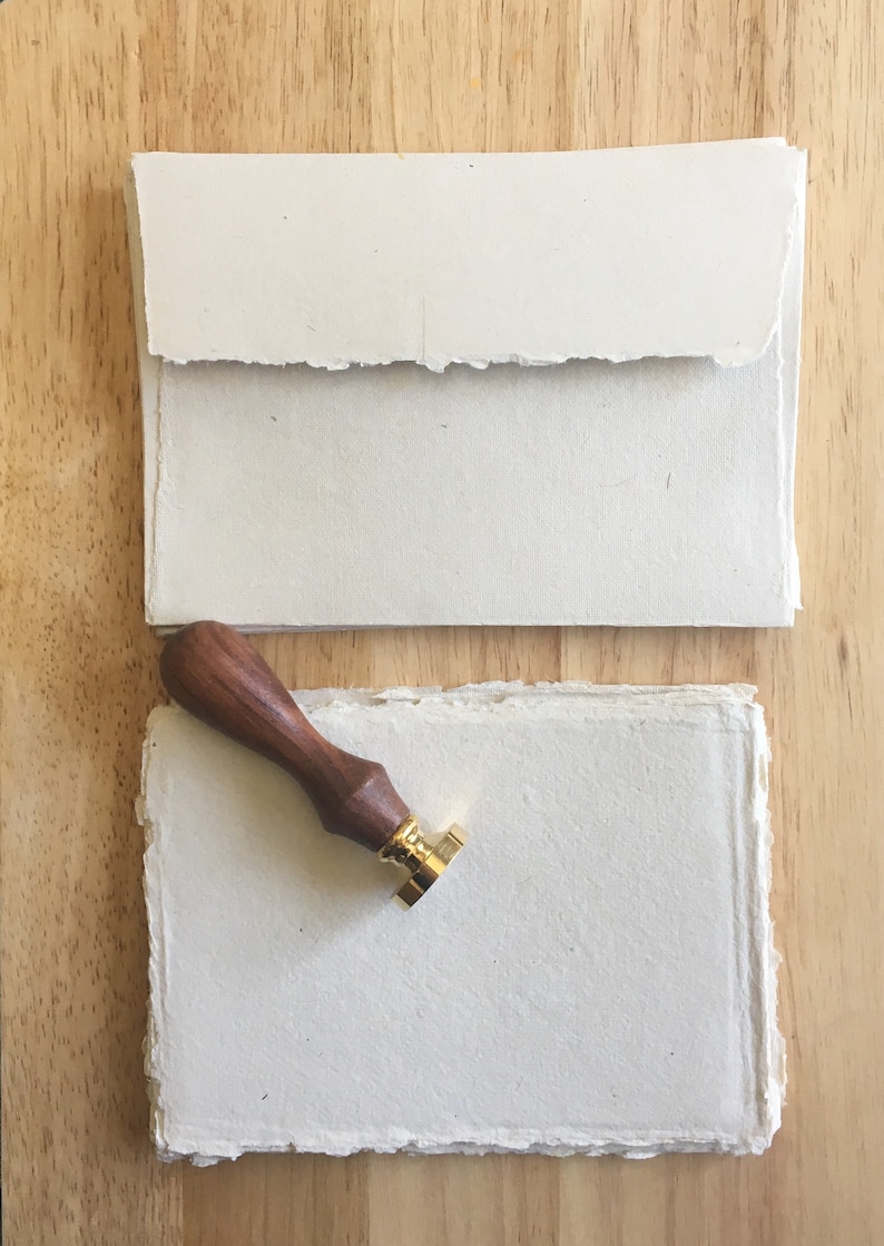 May include: A set of two blank, white, handmade paper sheets with deckled edges. A wooden stamp handle with a gold metal stamp head is on top of the sheets.