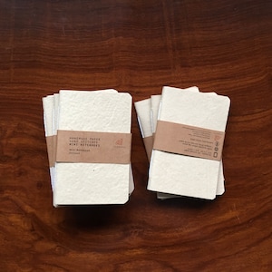 May include: Two stacks of handmade paper mini notebooks with brown paper covers. The cover of the stack on the left has the text "Handmade Paper Hand Stitched Mini Notebooks" and "Mini Notebook Unlined".